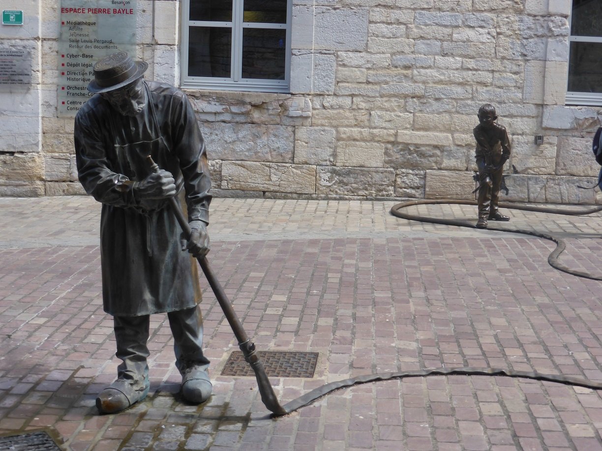the young boy has his foot on the hose and the old man is looking at the hose. Every so often water spurts into the mans face.